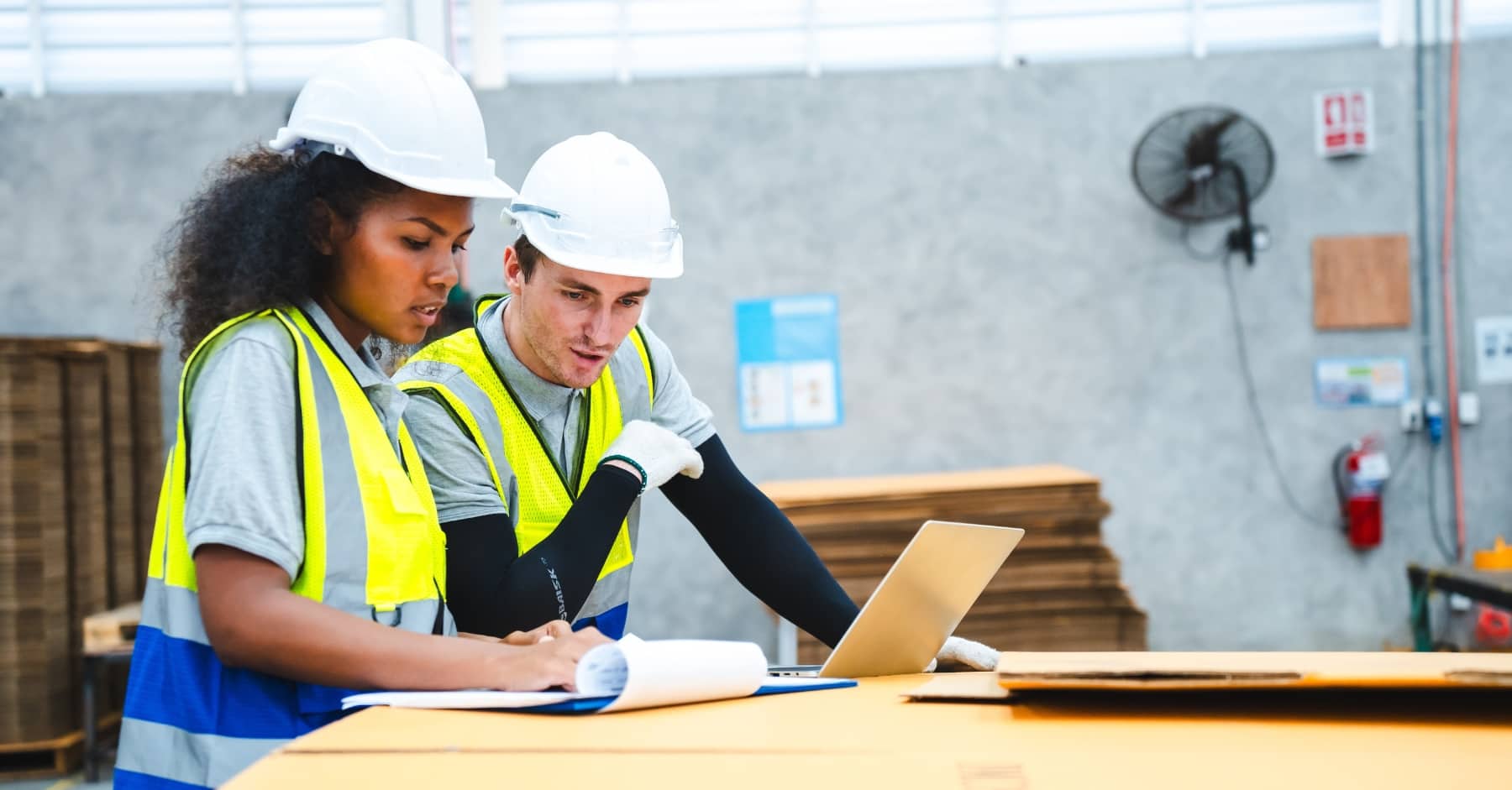 Two construction workers in hi-vis vests and hard hats reviewing documents and working on a laptop in a warehouse setting