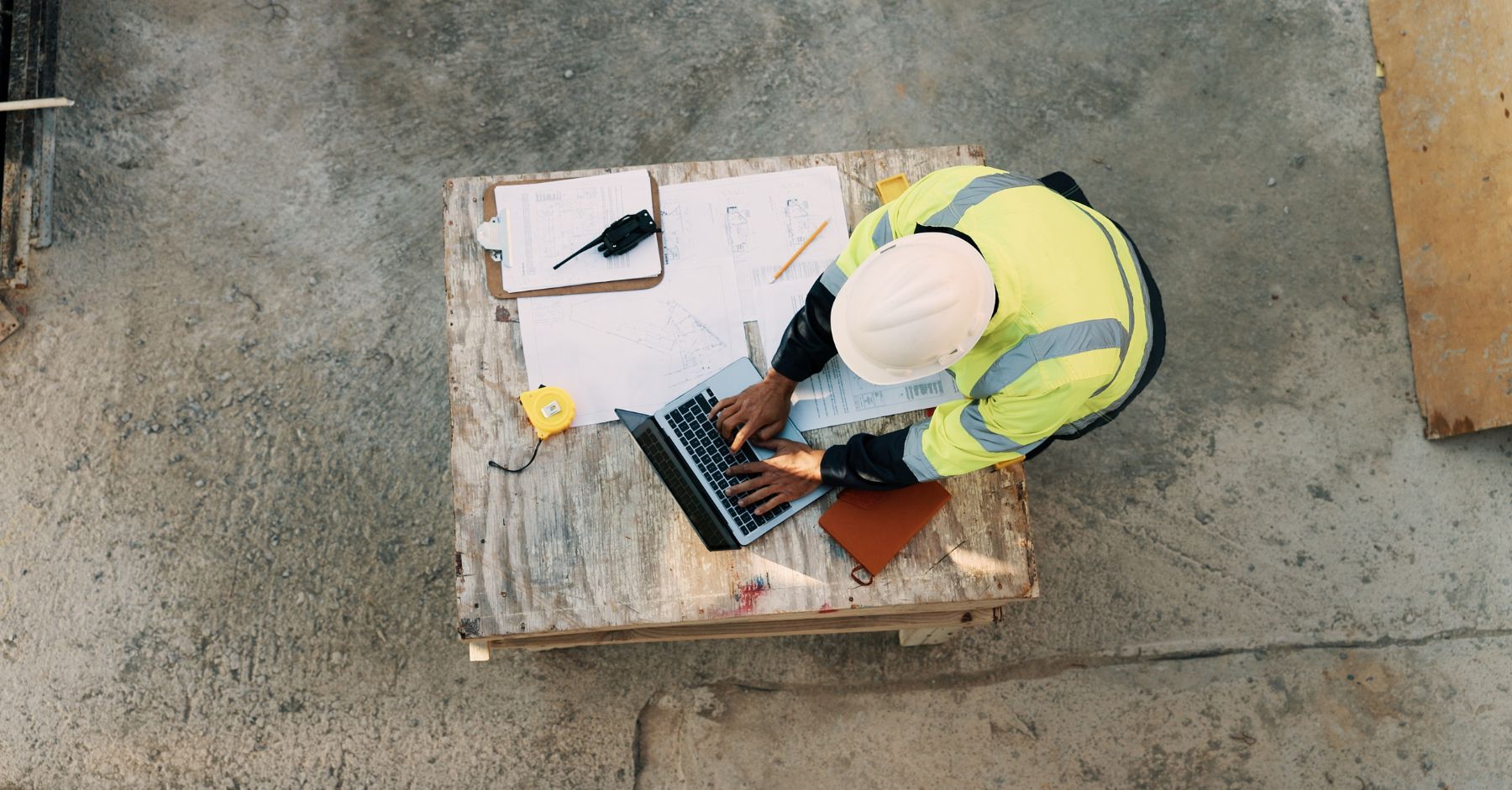 what does the autumn budget mean for construction jobs: An overhead view of a construction worker in a white hard hat and hi-vis jacket typing on a laptop at a wooden workbench on a concrete site, surrounded by building plans, a clipboard, a two-way radio and a yellow tape measure.