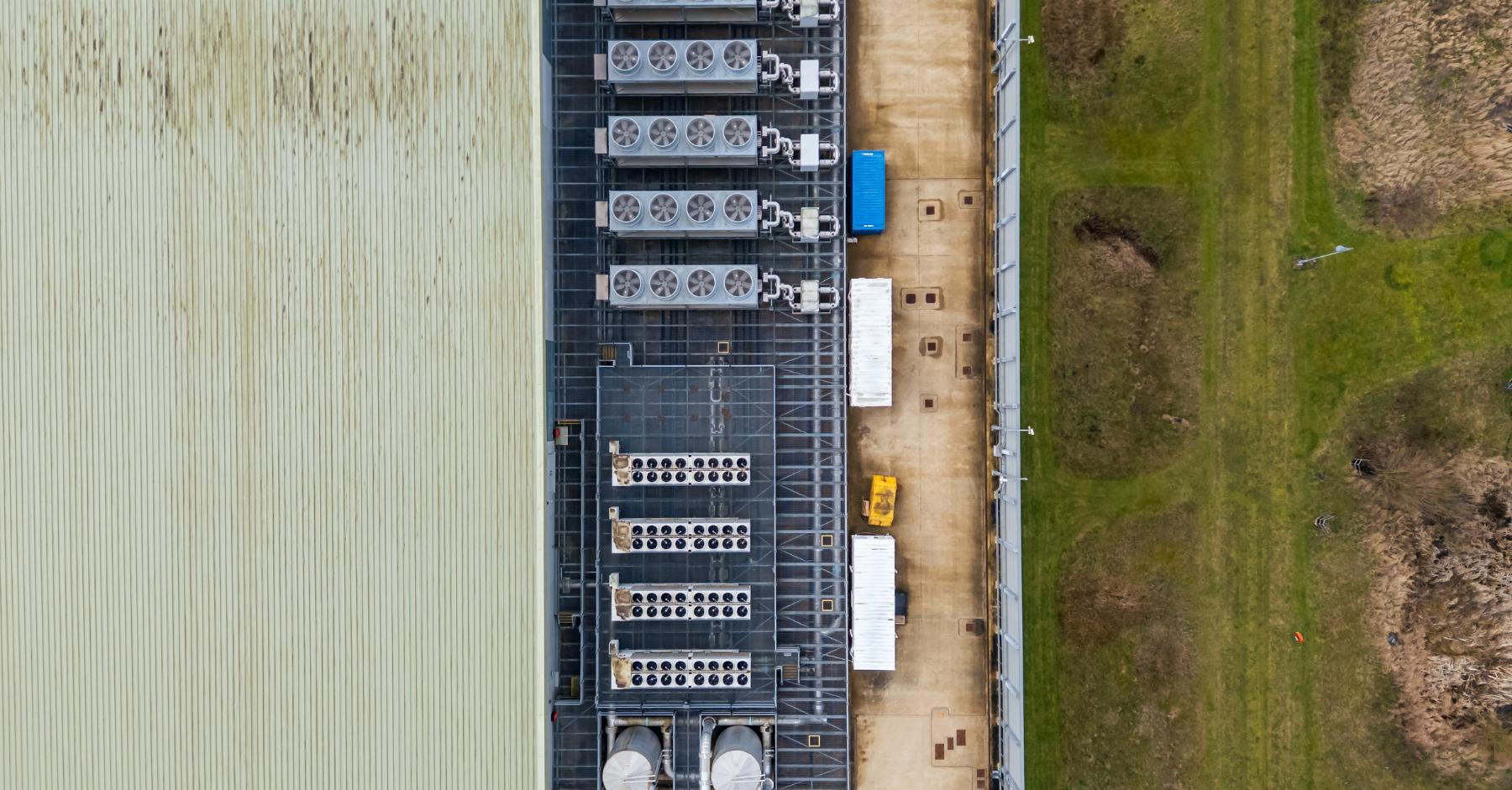 why work in industrial construction An aerial view of the roof and exterior of a large industrial warehouse building, with rows of HVAC units and ventilation equipment mounted along the roofline, flanked by a grassed area and a service road.